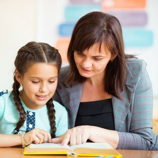 Female Teacher And Student Reading Book Together At Table