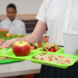 Female Student Holding Lunch Tray With Apple, Vegetables, Rice, And Beverage
