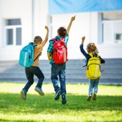 Three Students Jumping With Backpacks In Grass Infront Of School