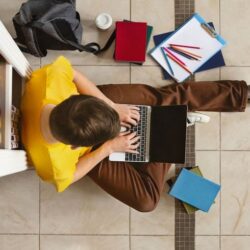Overhead View Of Male Student Sitting On Floor Using Laptop With Backpack C Coffee C Textbooks C And Pencils Around Him