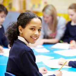 Female Student Of Color Wearing A Navy Blue Long Sleeve Smiling While Writing At Table