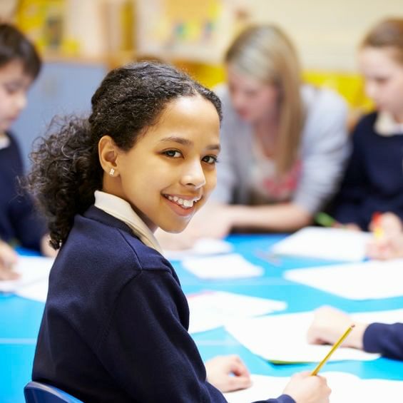 Female Student Of Color Wearing A Navy Blue Long Sleeve Smiling While Writing At Table