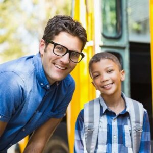 Teacher And Young Male Student Smiling At The Door Of A School Bus